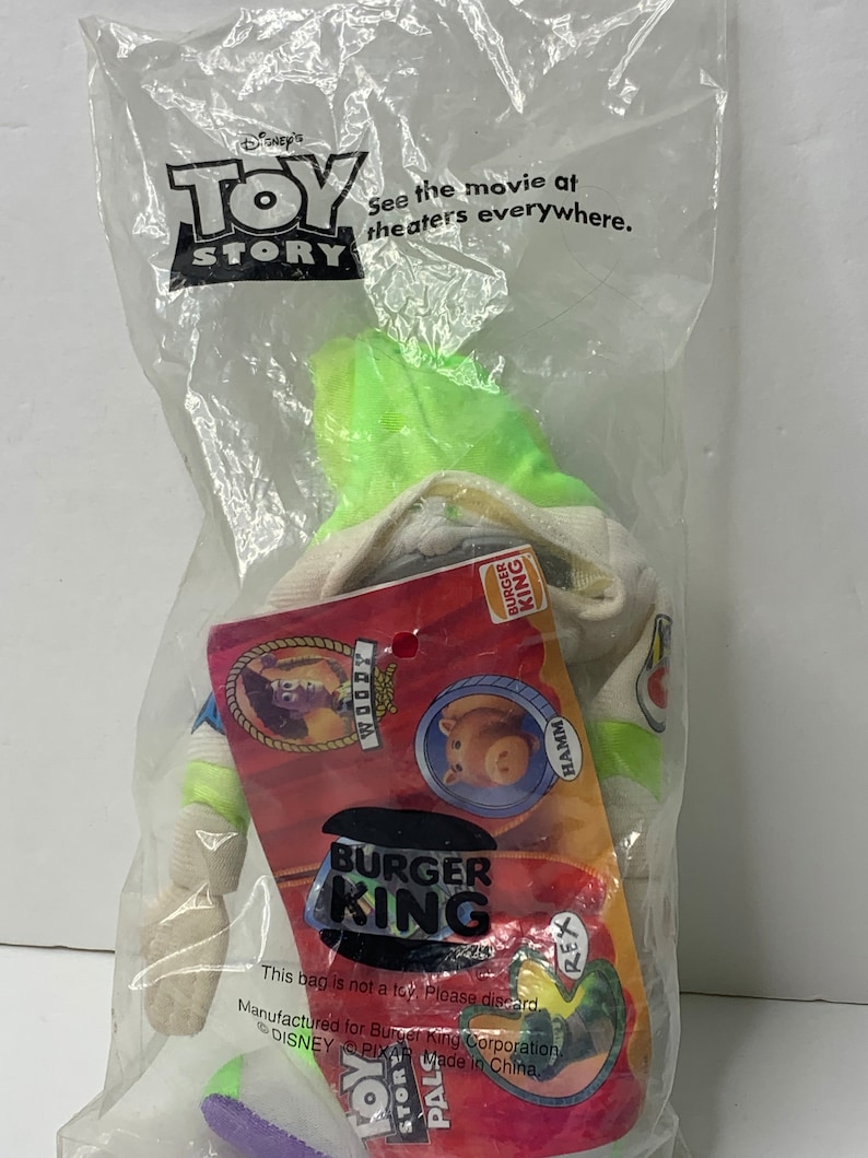 May include: A green Buzz Lightyear plush toy from the Disney Pixar movie Toy Story. The toy is wearing a Burger King uniform with the Burger King logo and the words "See the movie at theaters everywhere." The toy is packaged in a clear plastic bag with a Burger King logo.