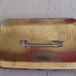 May include: A rectangular pinback button with a safety pin clasp on the back. The button has a metallic gold surface with rounded corners. The edges of the button have a colorful design.