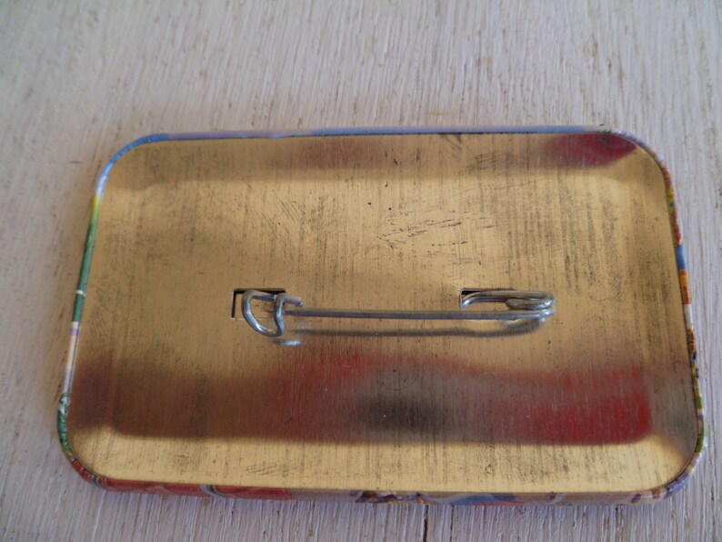May include: A rectangular pinback button with a safety pin clasp on the back. The button has a metallic gold surface with rounded corners. The edges of the button have a colorful design.