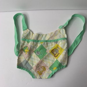 Pode incluir: Um porta-bebê de boneca verde e branco com um padrão xadrez e imagens de bonecas segurando balões. O porta-bebê tem duas alças verdes.