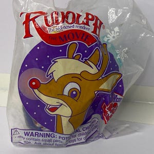 May include: Sealed plastic package featuring a Rudolph the Red-Nosed Reindeer toy. The package has the movie title in red and the cartoon reindeer on a purple background. The package includes a warning about small parts.