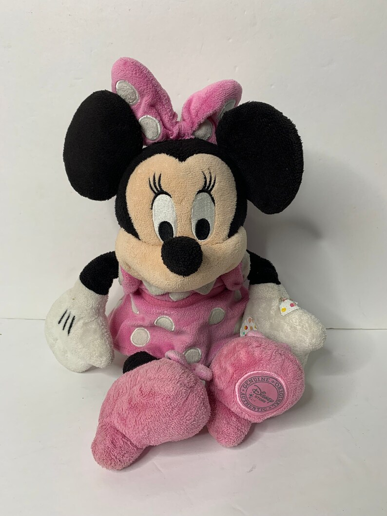 May include: A pink and white polka dot Minnie Mouse plush toy with a pink bow. The toy is sitting with its legs crossed and has a white face with black eyes and eyelashes. The toy has a pink and white polka dot dress and a white tag that says "Disney Genuine Original".