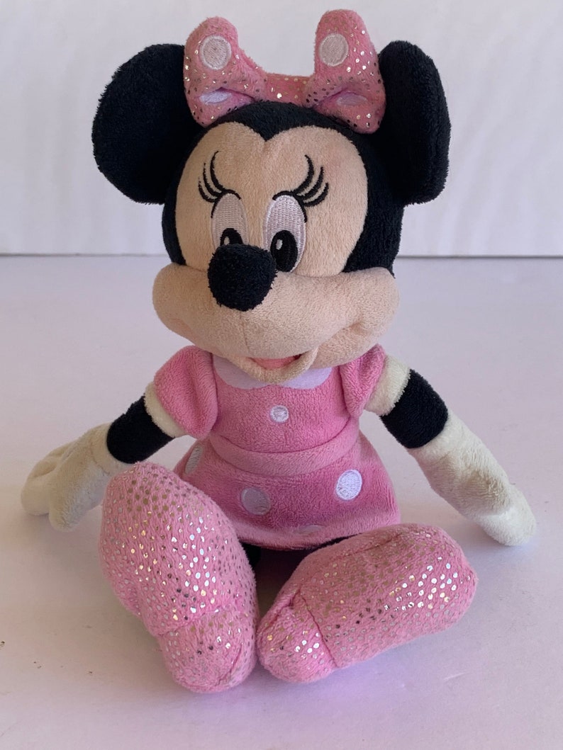 May include: A pink plush Minnie Mouse toy with black ears, a pink bow with white polka dots, and a pink dress with white polka dots. The toy is sitting with its legs crossed and has a sparkly pink finish.