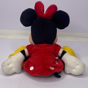 Pu&ograve; includere: Una bambola di peluche di Minnie Mouse, vista da dietro, con orecchie nere, un fiocco rosso e un vestito rosso a pois bianchi. La bambola ha guanti bianchi e maniche gialle. La bambola &egrave; seduta.