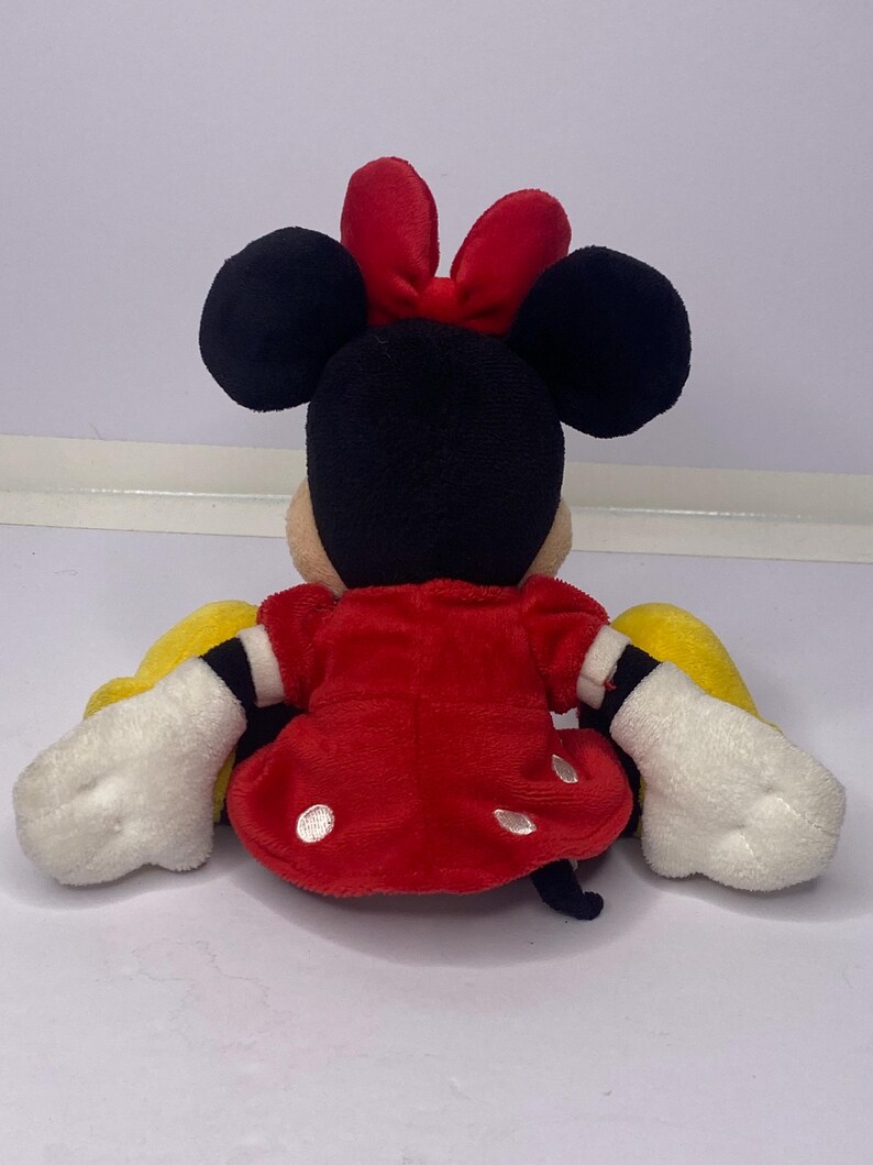 Pu&ograve; includere: Una bambola di peluche di Minnie Mouse, vista da dietro, con orecchie nere, un fiocco rosso e un vestito rosso a pois bianchi. La bambola ha guanti bianchi e maniche gialle. La bambola &egrave; seduta.