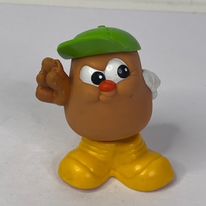 Op de afbeelding: Een vintage Mr. Potato Head speelgoedfiguur. Het speelgoed heeft een bruine kop, witte ogen, een rode neus en een groene pet. Het draagt gele schoenen en heeft een hand in een gebaar geheven. Een klassiek speelgoed.