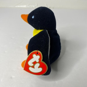 Puede incluir: Un peluche de pingüino beanie baby negro y amarillo con una etiqueta de corazón rojo Ty. El pingüino está sentado sobre una superficie blanca.