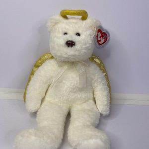 K&ouml;nnte beinhalten: Ein wei&szlig;er Teddyb&auml;r mit goldenem Heiligenschein und goldenen Glitzerfl&uuml;geln. Der B&auml;r hat eine braune Nase und kleine schwarze Augen. Ein rotes herzf&ouml;rmiges Etikett ist am Ohr befestigt. Ein wei&szlig;es Band ist um den Hals gebunden.
