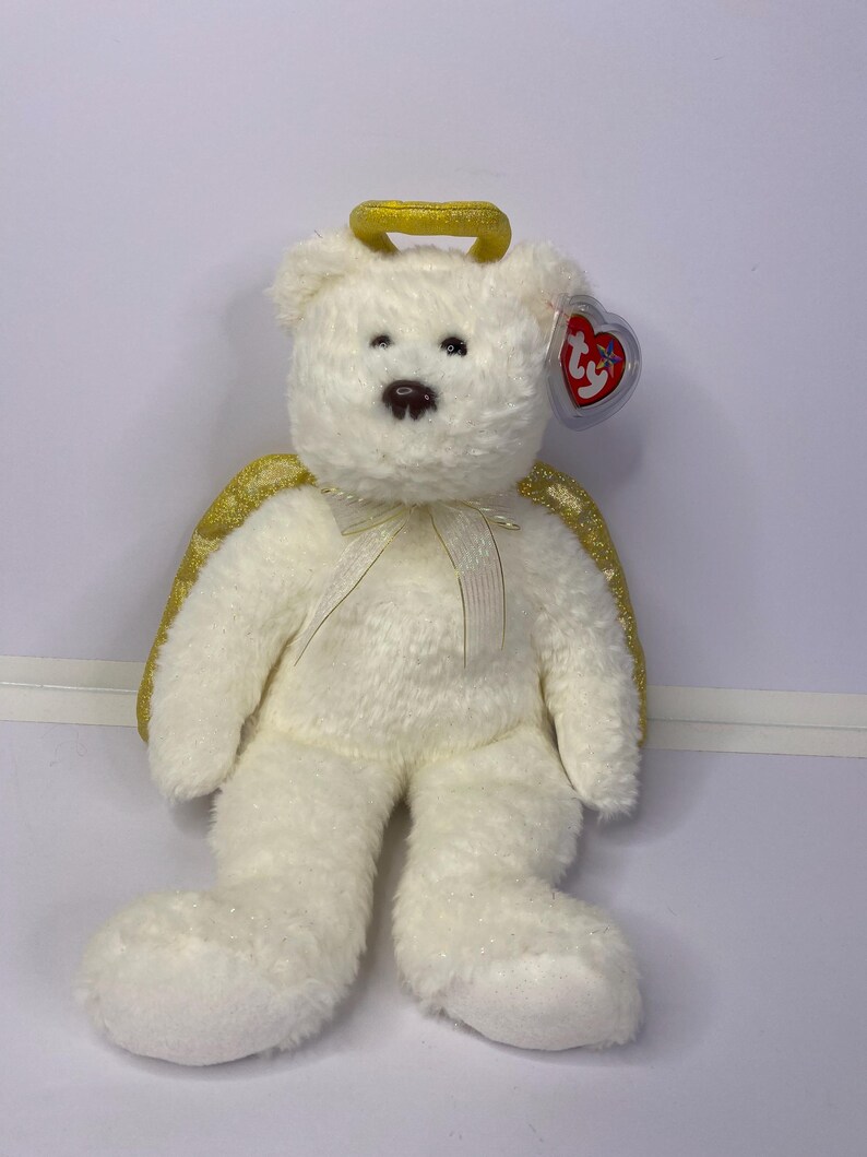 K&ouml;nnte beinhalten: Ein wei&szlig;er Teddyb&auml;r mit goldenem Heiligenschein und goldenen Glitzerfl&uuml;geln. Der B&auml;r hat eine braune Nase und kleine schwarze Augen. Ein rotes herzf&ouml;rmiges Etikett ist am Ohr befestigt. Ein wei&szlig;es Band ist um den Hals gebunden.