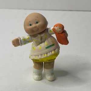 Op de afbeelding: Een Cabbage Patch Kid pop die een geel en wit jurkje draagt met een citroenprint. De pop houdt een kleine rode pop vast met een geel jurkje.