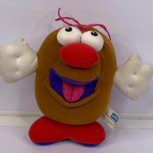 Op de afbeelding: Een pluche speelgoed in de vorm van een aardappel met grote witte ogen, een rode neus en een paarse mond met een roze tong. Het heeft witte handen, rode voeten en een bruin lichaam. Roze touw is aan de bovenkant bevestigd.