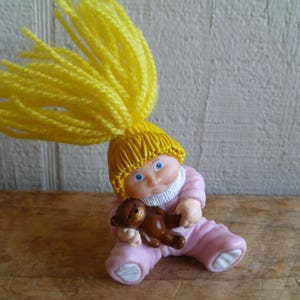 Op de afbeelding: Een kleine plastic pop met geel garen haar, die een roze outfit draagt en een bruine teddybeer vasthoudt.