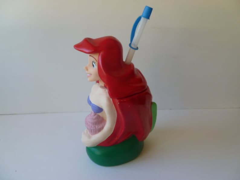 May include: Red and green plastic cup shaped like Ariel, the Little Mermaid, with a blue straw. The cup has a shell design and a purple top.