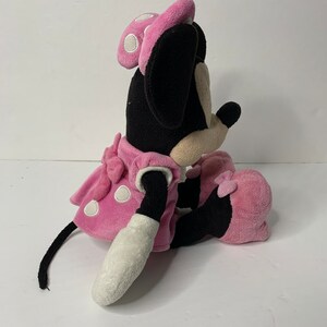 May include: A pink plush Minnie Mouse toy with white polka dots on her dress and bow. She is sitting with her legs crossed and her tail is curled behind her.