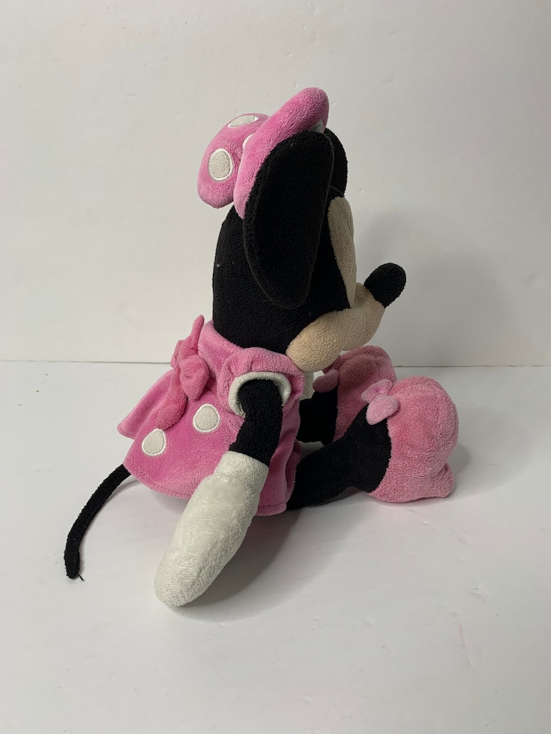 May include: A pink plush Minnie Mouse toy with white polka dots on her dress and bow. She is sitting with her legs crossed and her tail is curled behind her.