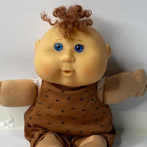 Könnte beinhalten: Eine Cabbage Patch Kid Puppe mit braunen Locken, blauen Augen und einem braunen und weißen gemusterten Outfit. Die Puppe sitzt mit gekreuzten Beinen.