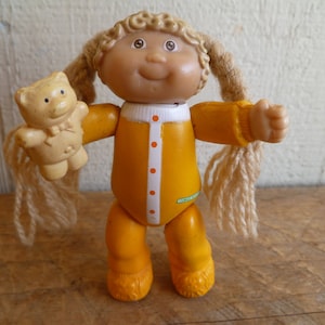 Op de afbeelding: Een Cabbage Patch Kid pop gekleed in geel met blond garen haar houdt een kleine, crèmekleurige teddybeer vast. De pop heeft een geschilderd gezicht met blauwe ogen en een lieve glimlach.
