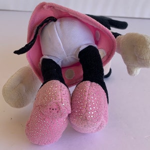 May include: Pink and black plush toy with white polka dots. The toy is wearing a pink dress with white polka dots and has a black tail. The toy is sitting on a white surface.