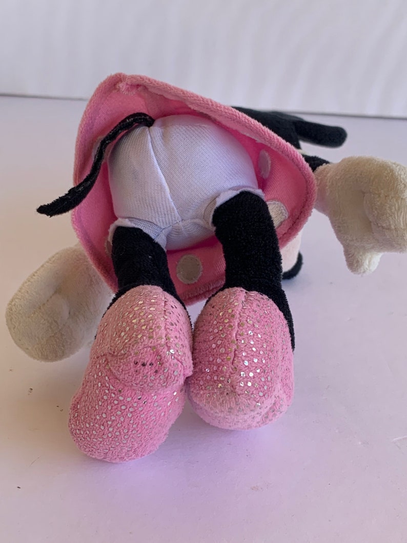 May include: Pink and black plush toy with white polka dots. The toy is wearing a pink dress with white polka dots and has a black tail. The toy is sitting on a white surface.