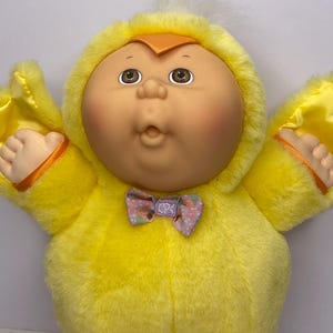 Pode incluir: Uma boneca Cabbage Patch Kids vintage vestida com uma fantasia de pintinho amarelo. A boneca tem um rosto redondo, olhos castanhos e bochechas rosadas. A fantasia apresenta um capuz, asas e uma gravata borboleta com as iniciais "CPK".