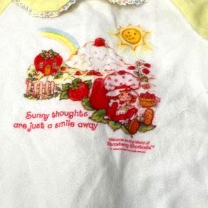 May include: White and yellow Strawberry Shortcake themed onesie. The onesie features a graphic of Strawberry Shortcake, a rainbow, and the text "Sunny thoughts are just a smile away". The collar and sleeves are yellow.