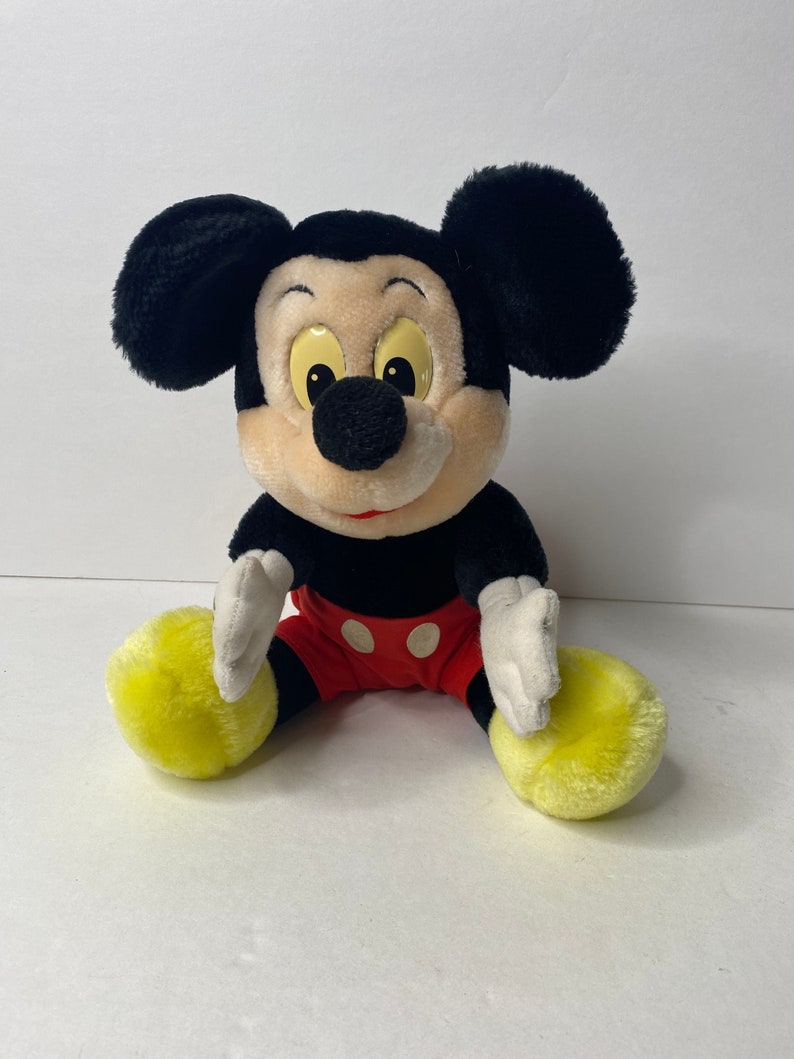 May include: A plush Mickey Mouse toy sitting with his legs crossed. He is wearing red shorts with white polka dots and yellow shoes. His ears are black and his face is brown with black eyes and a black nose.