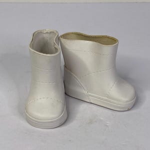 May include: A pair of white ankle boots featuring a smooth finish, a rounded toe, and subtle stitched detailing along the sides. The boots have a flat sole and a structured shaft, with one boot standing upright and the other positioned on its side.