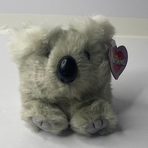 May include: A gray plush koala bear sitting with its paws in front of it. The bear has a black nose and a white tag that says "Puffins" with a red heart.