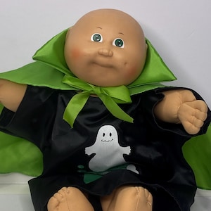 Könnte beinhalten: Cabbage Patch Kid Puppe in einem schwarzen Satin-Kostüm mit einem weißen Geister-Motiv. Das Kostüm hat einen grünen Satin-Kragen und eine Schleife. Die Puppe hat einen kahlen Kopf, rosige Wangen und grüne Augen.