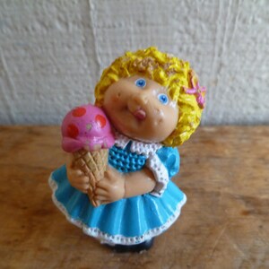 May include: A small plastic doll with blonde hair wearing a blue dress and holding a pink and white ice cream cone.
