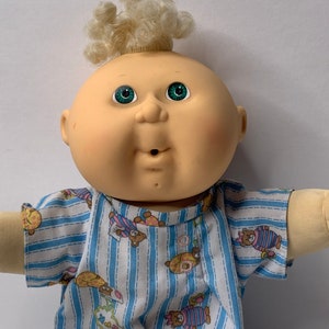 May include: A Cabbage Patch Kid doll with blonde curly hair, green eyes, and a pink and blue striped shirt with teddy bear designs.