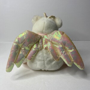 May include: White plush angel bear with iridescent wings. The bear has a gold halo and is sitting with its wings spread out.