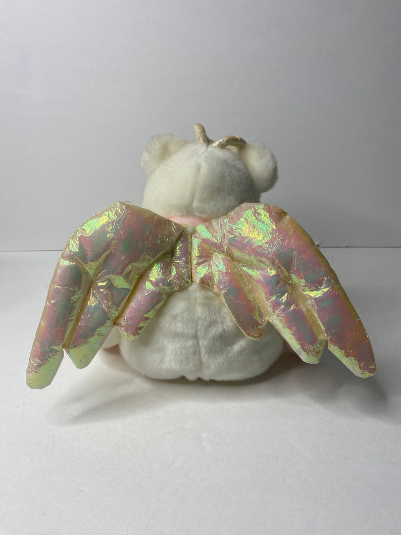 May include: White plush angel bear with iridescent wings. The bear has a gold halo and is sitting with its wings spread out.