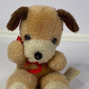 Puede incluir: Un juguete de peluche vintage con forma de perro. Tiene un cuerpo marrón, patas y hocico blancos y orejas marrones. El juguete tiene una nariz negra, ojos negros y una cinta roja. Una etiqueta está adjunta.