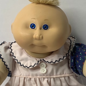 Puede incluir: Una muñeca Cabbage Patch Kid con cabello rubio, ojos azules y un vestido rosa con cuello blanco y botones. La muñeca lleva una camisa de estampado azul y blanco debajo del vestido.