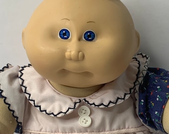 Puede incluir: Una muñeca Cabbage Patch Kid con cabello rubio, ojos azules y un vestido rosa con cuello blanco y botones. La muñeca lleva una camisa de estampado azul y blanco debajo del vestido.