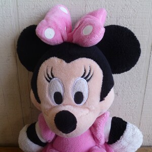 Puede incluir: Un juguete de peluche de un rat&oacute;n de dibujos animados con grandes orejas negras, un lazo rosa con lunares blancos y ojos negros. El rat&oacute;n lleva un vestido rosa con ribetes blancos.