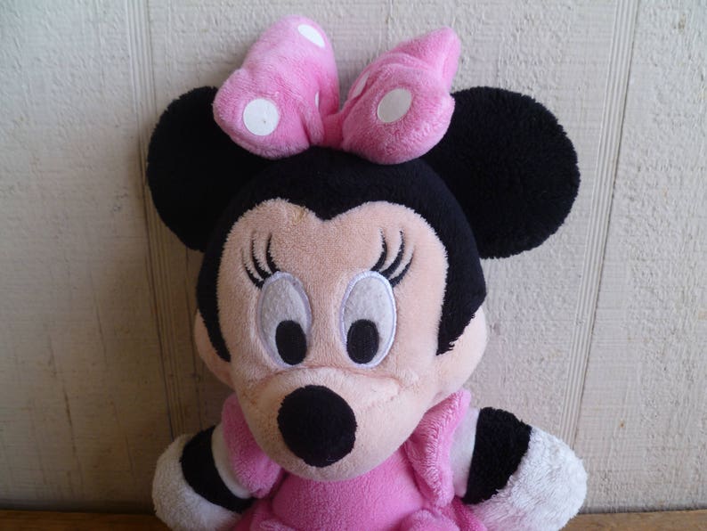 Puede incluir: Un juguete de peluche de un rat&oacute;n de dibujos animados con grandes orejas negras, un lazo rosa con lunares blancos y ojos negros. El rat&oacute;n lleva un vestido rosa con ribetes blancos.