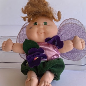 May include: A Cabbage Patch Kid doll dressed as a fairy with purple flowers and green leaves. The doll has brown hair, green eyes, and a pink dress. The wings are purple and have a sparkly trim.