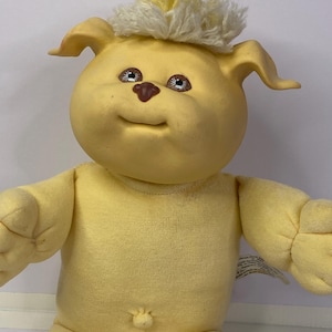 Peut inclure: Une peluche jaune vintage en forme de créature ressemblant à un chien. Elle a une tête ronde, des oreilles tombantes et un petit nez marron. Le jouet a une touffe de cheveux jaunes sur le dessus de la tête et porte une tenue jaune.