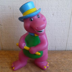 May include: A purple dinosaur toy wearing a blue and yellow top hat and a green vest. The dinosaur is holding a yellow and green stick in its hands.