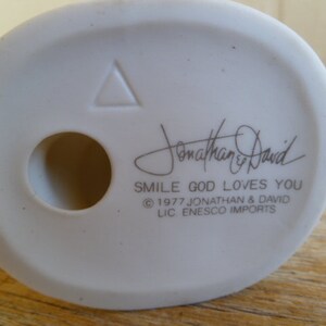 Op de afbeelding: Witte keramische figuur met een driehoek en een gat in de bodem. De figuur heeft de tekst "Jonathan & David" en "Smile God Loves You" met het copyright-symbool en "1977 Jonathan & David LIC. ENESCO IMPORTS".