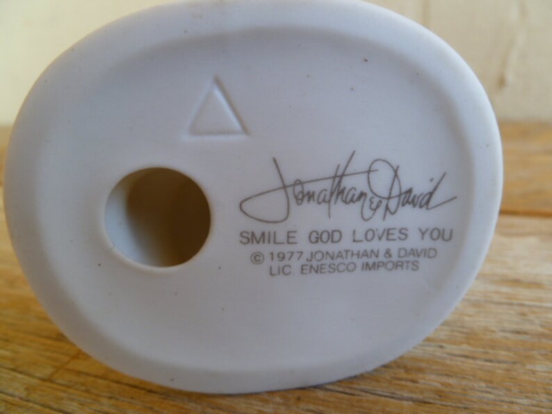 Op de afbeelding: Witte keramische figuur met een driehoek en een gat in de bodem. De figuur heeft de tekst "Jonathan & David" en "Smile God Loves You" met het copyright-symbool en "1977 Jonathan & David LIC. ENESCO IMPORTS".