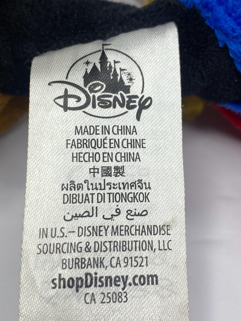 May include: A white tag with the Disney logo and text in multiple languages, including "MADE IN CHINA" and "shopDisney.com". The tag also includes the address of Disney Merchandise Sourcing & Distribution, LLC in Burbank, CA.