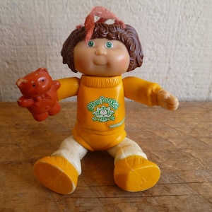 Op de afbeelding: Een Cabbage Patch Kids pop die een geel shirt draagt met een groen logo waarop "Cabbage Patch Kids" staat. De pop houdt een bruine teddybeer vast en heeft bruin haar en blauwe ogen.