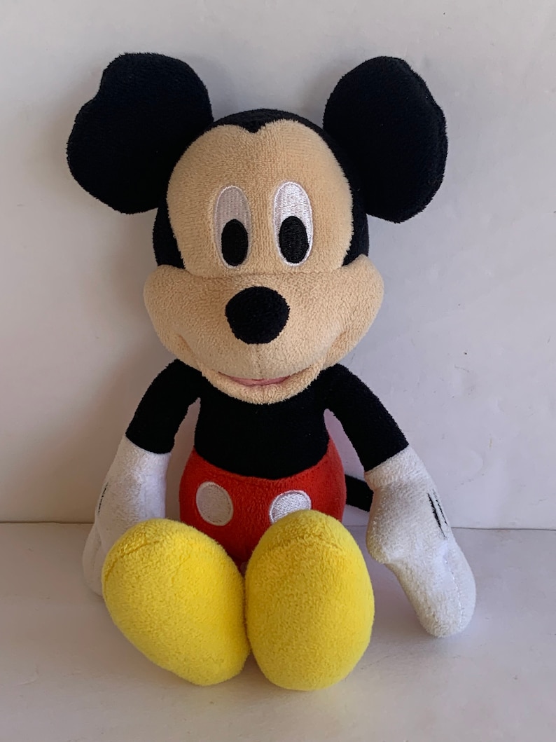 Op de afbeelding: Een pluche Mickey Mouse-speelgoed. Het speelgoed heeft een zwarte kop met grote, ronde oren, een beige gezicht en wit omrande ogen. Het draagt een zwarte torso, rode shorts, witte handschoenen en gele voeten. Het speelgoed zit rechtop.