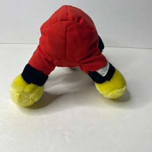 May include: A plush toy of Mickey Mouse in a red and black outfit. The toy is sitting on a white surface.