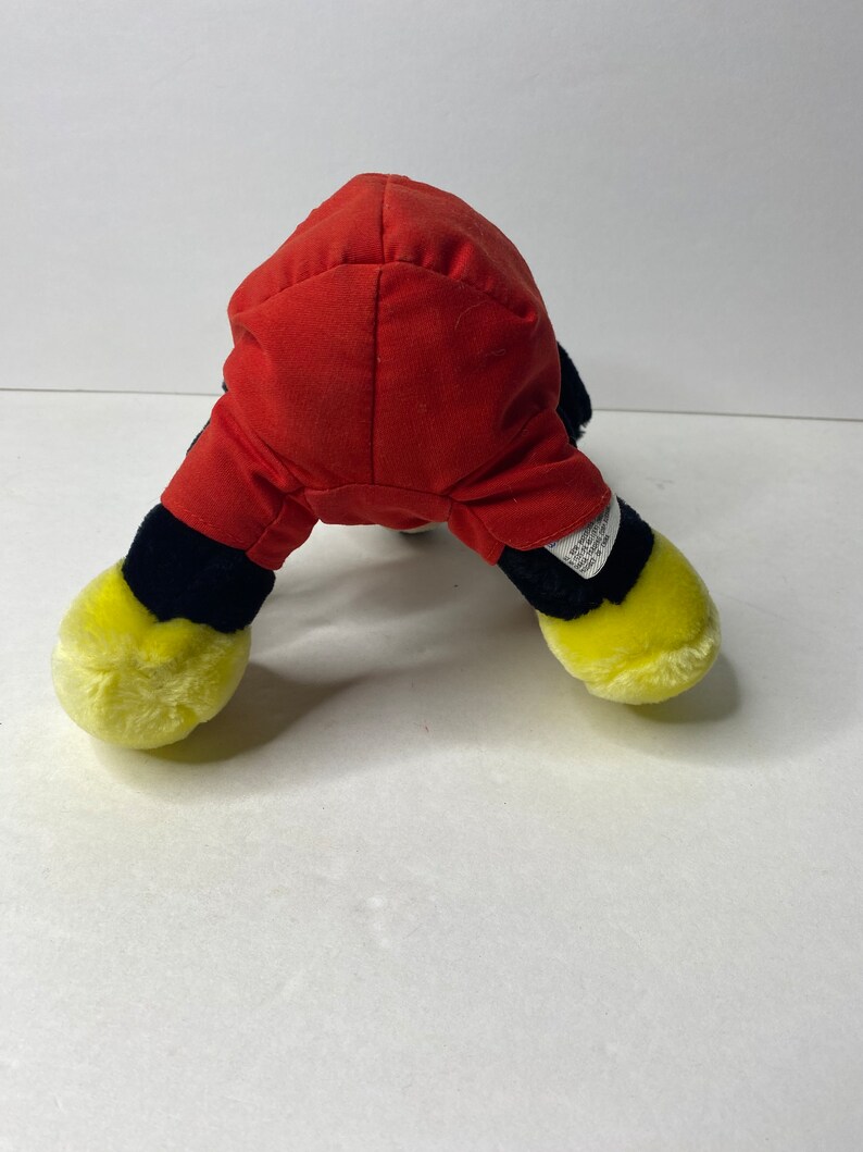 May include: A plush toy of Mickey Mouse in a red and black outfit. The toy is sitting on a white surface.