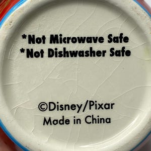 Puede incluir: Primer plano del interior inferior de una taza de cer&aacute;mica. El texto dice "*Not Microwave Safe *Not Dishwasher Safe &copy;Disney/Pixar Made in China". La taza tiene un interior blanco con un esmalte agrietado.