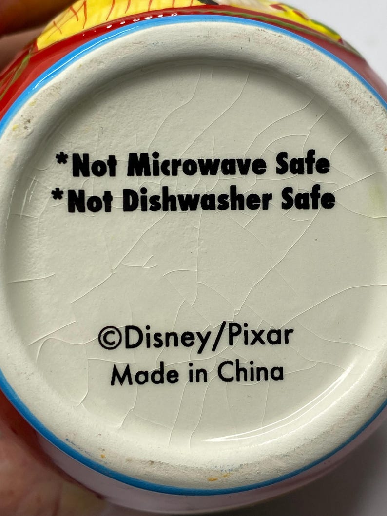 Puede incluir: Primer plano del interior inferior de una taza de cer&aacute;mica. El texto dice "*Not Microwave Safe *Not Dishwasher Safe &copy;Disney/Pixar Made in China". La taza tiene un interior blanco con un esmalte agrietado.
